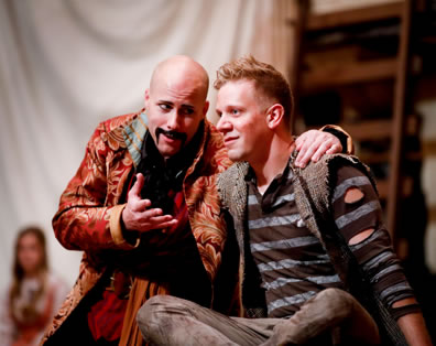 Stache, bald head, thick (fake) fu manchu mustache around his lips an wearing a tapestry-like gold coat has his left arm around Peter, who's wearing a holy striped shirt, grubby jeans, and burlap vest, as they confer. In the background is the actress playing Molly sitting, watching, in front of a white drape