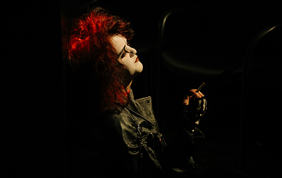 First Witch looks up with bright red hair and stark makeup, wearing a black leather jacket, and a cigeratte in her fingerless-gloved right hand.